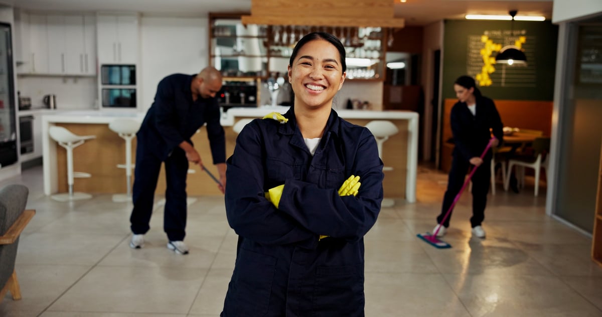 Happy female janitor in uniform smiling with arms crossed, team in background